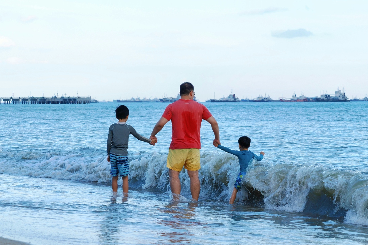 An adult in a red shirt and yellow swim trunks stands in the surf holding the hands of two young boys in rash guards as a wave breaks around them, all three photographed from behind. A industrial harbor with cargo ships and a pier stretches across the horizon under an overcast sky.