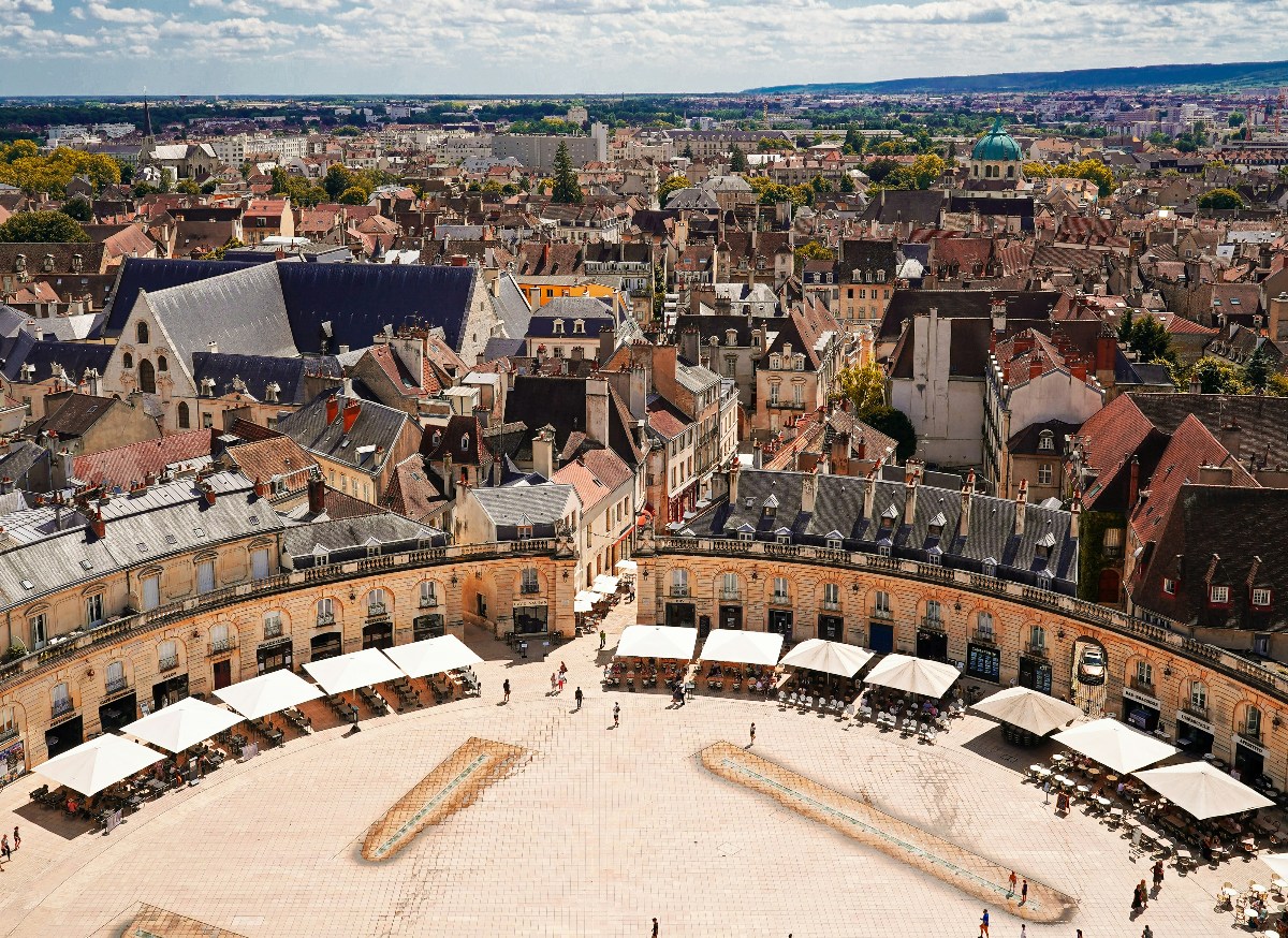 An aerial view looks down onto a large semicircular stone plaza lined with café terraces shaded by white umbrellas, with small figures of pedestrians crossing the open paved center. Beyond the curved classical façade, a dense cityscape of cream stone and terracotta-roofed buildings extends to the horizon, with a green-domed church visible among the rooftops.