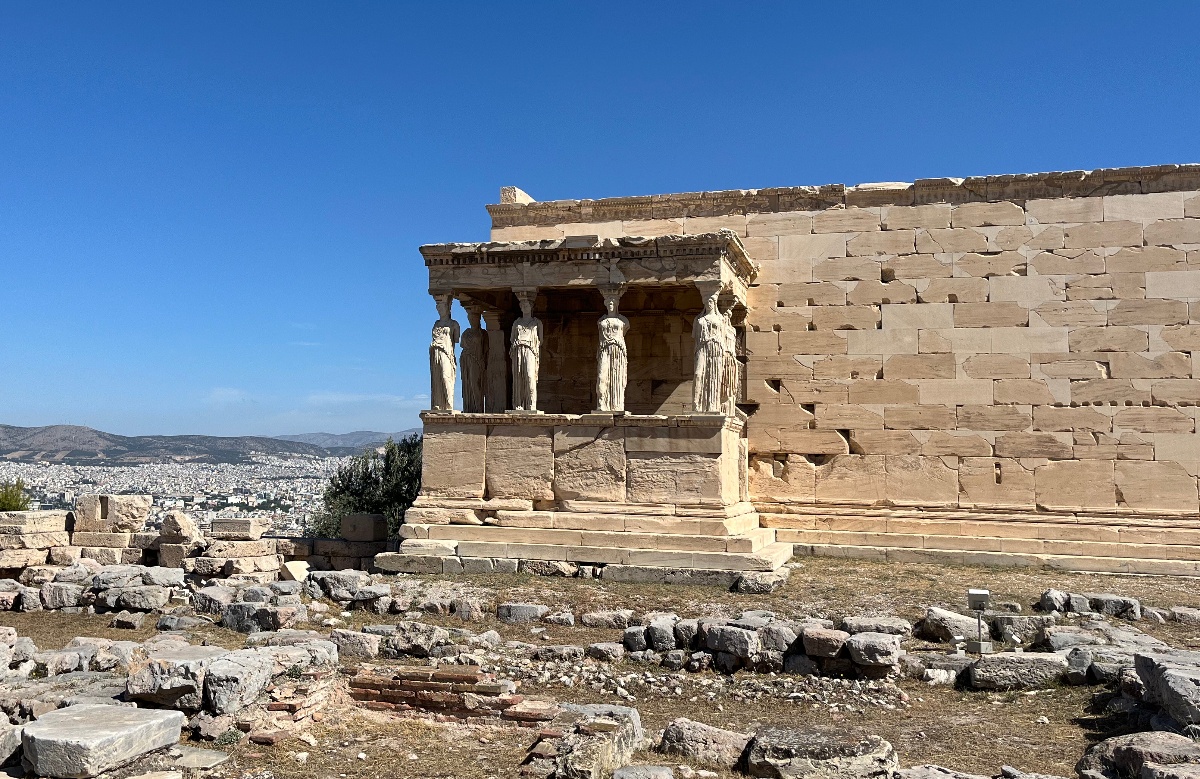 The Erechtheion's Porch of the Caryatids, where six draped female statues serve as columns supporting the roof, projects from the side of the marble temple on the Acropolis. Scattered stone ruins lie in the foreground, with the city of Athens and distant hills visible beyond the hilltop under a clear blue sky.