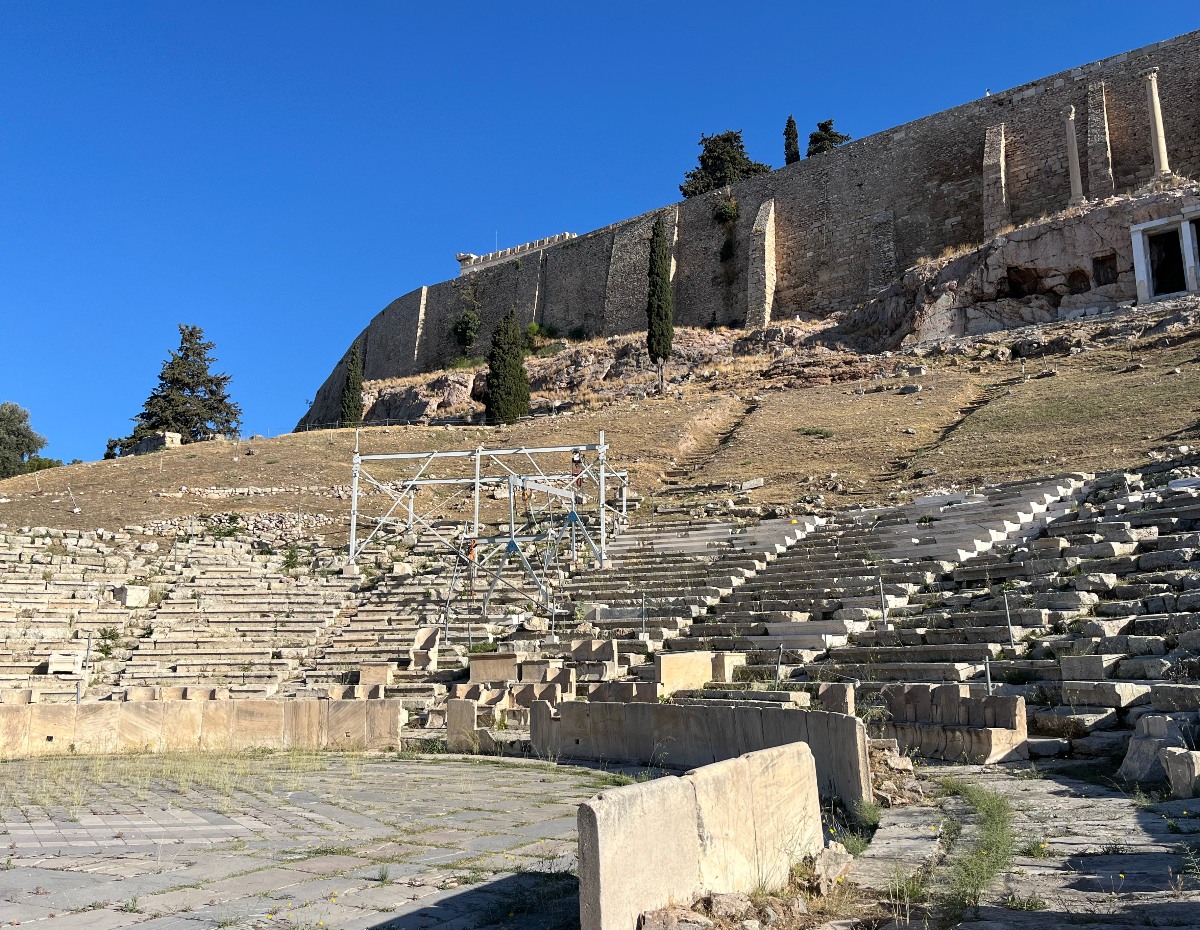 The Theatre of Dionysus, with its weathered stone seating tiers rising up a hillside, sits below the fortified southern wall of the Acropolis. Metal scaffolding stands among the ruined rows, while cypress trees and dry grass cover the slope beneath the ancient citadel under a clear blue sky.