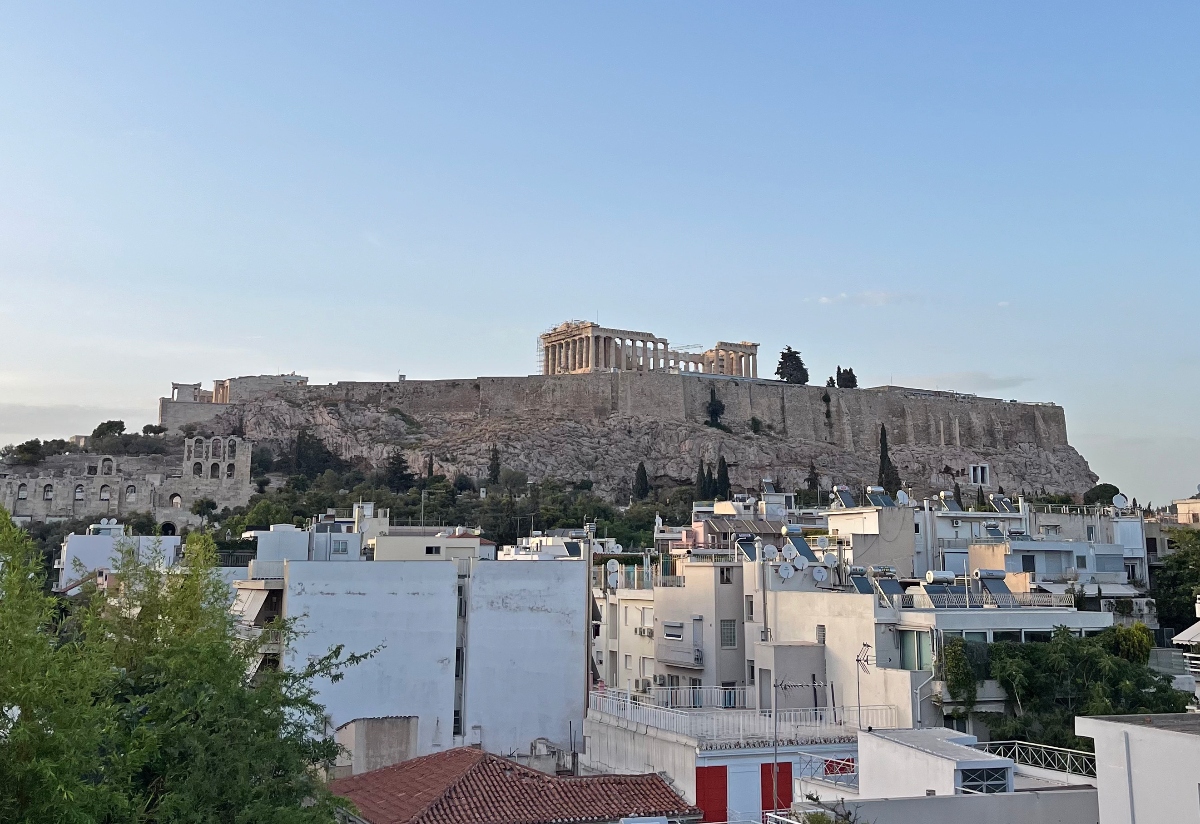 The Acropolis of Athens rises on a rocky hill, with the columned Parthenon visible at its summit and the arched stone facade of the Odeon of Herodes Atticus at its lower left. In the foreground, white residential buildings with rooftop solar water heaters and satellite dishes sit below the ancient site under a clear pale-blue sky.