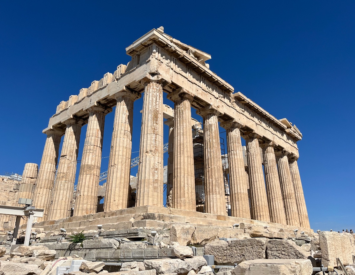 The Parthenon, viewed from a corner showing two colonnaded sides of fluted Doric columns supporting a triangular pediment and entablature, stands against a deep blue sky on the Acropolis. Scattered marble blocks and scaffolding from ongoing restoration work lie around the temple's base.