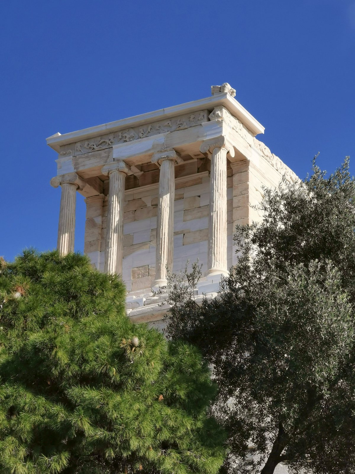 The Temple of Athena Nike, a small white marble structure with fluted Ionic columns and a carved frieze, stands against a deep blue sky on the Acropolis. Green pine and olive trees frame the lower portion of the temple in the foreground.