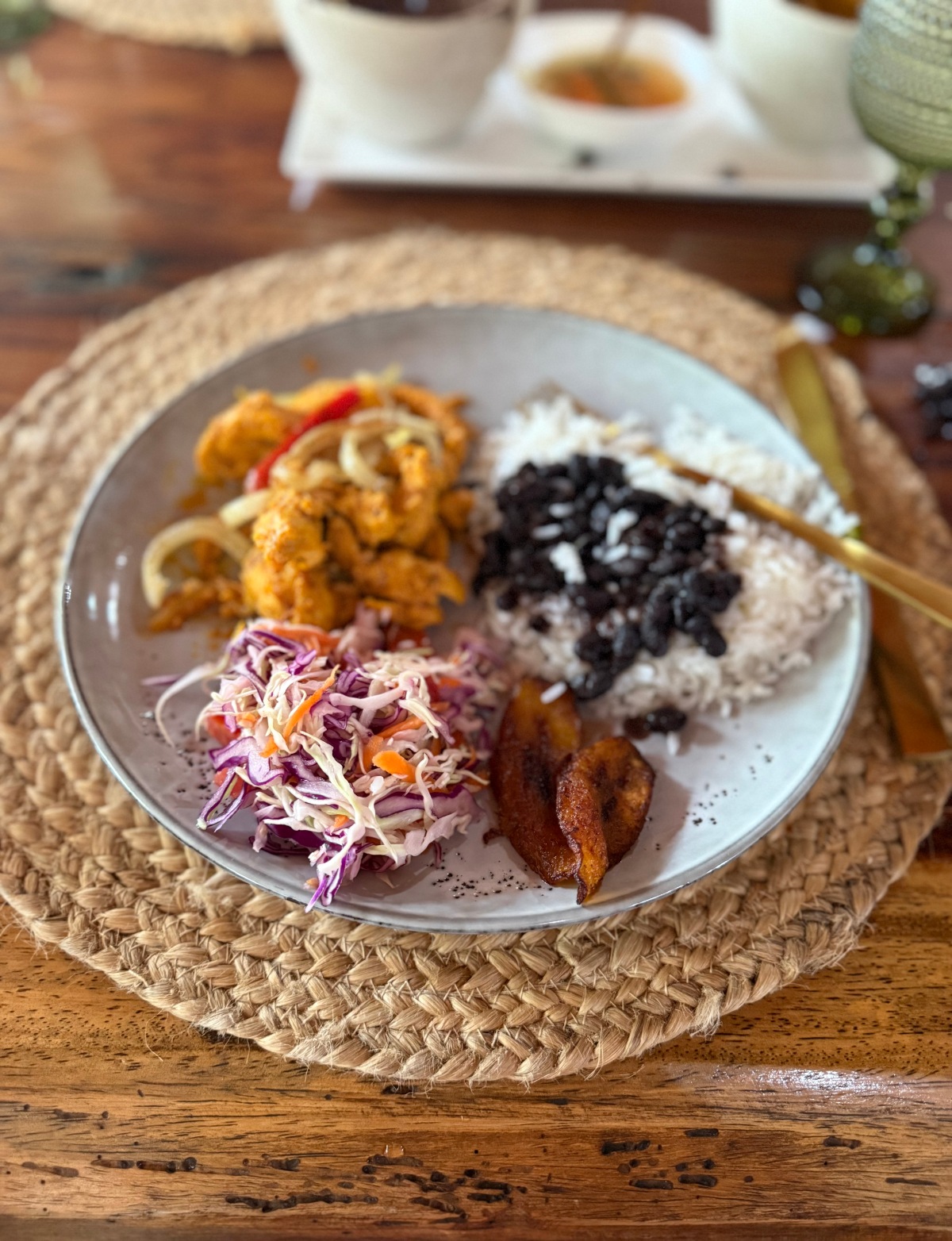 A white plate holds a traditional Costa Rican casado with white rice topped with black beans, seasoned chicken with sautéed onions and red peppers, fried plantain slices, and a shredded cabbage and carrot slaw. The plate sits on a woven jute placemat atop a wooden table, with a gold fork resting on its edge.
