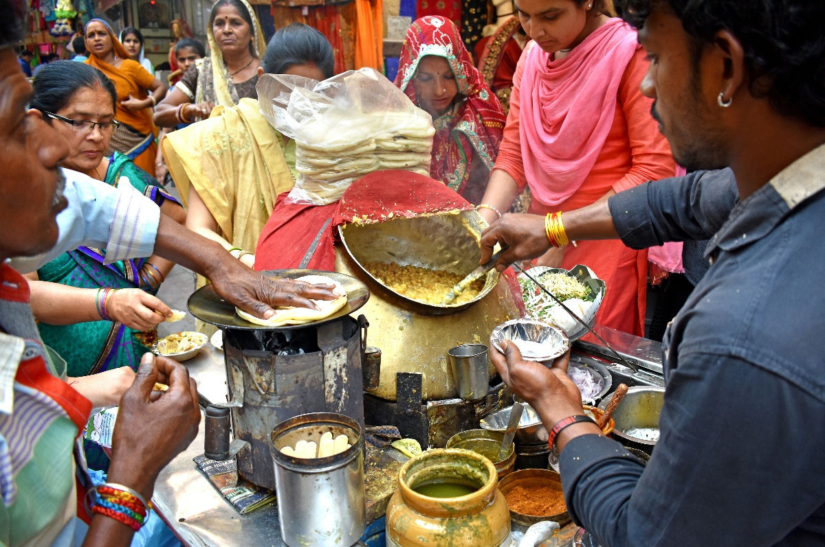 A vendor serves chickpea curry from a large pot at a street food stall while a stack of flatbreads sits wrapped in plastic nearby, and a customer's hand reaches toward a plate of food on a griddle. Several women in colorful saris and a man wait around the stall amid pots, bowls, and other cooking equipment.