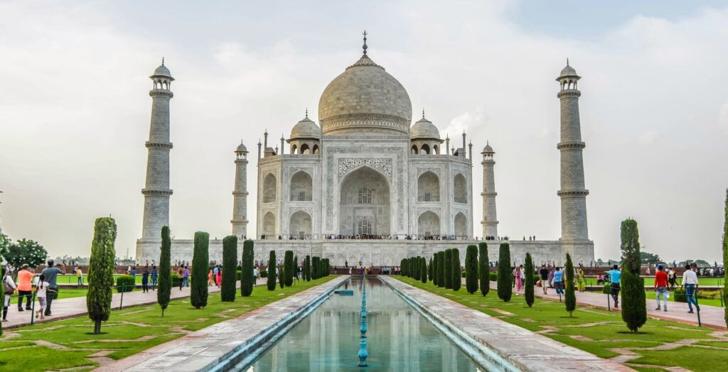 The Taj Mahal's white marble mausoleum, topped by a central dome and flanked by four minarets, is reflected in a long rectangular pool lined with cypress trees and manicured lawns. Visitors walk along the surrounding pathways and gardens beneath an overcast sky.