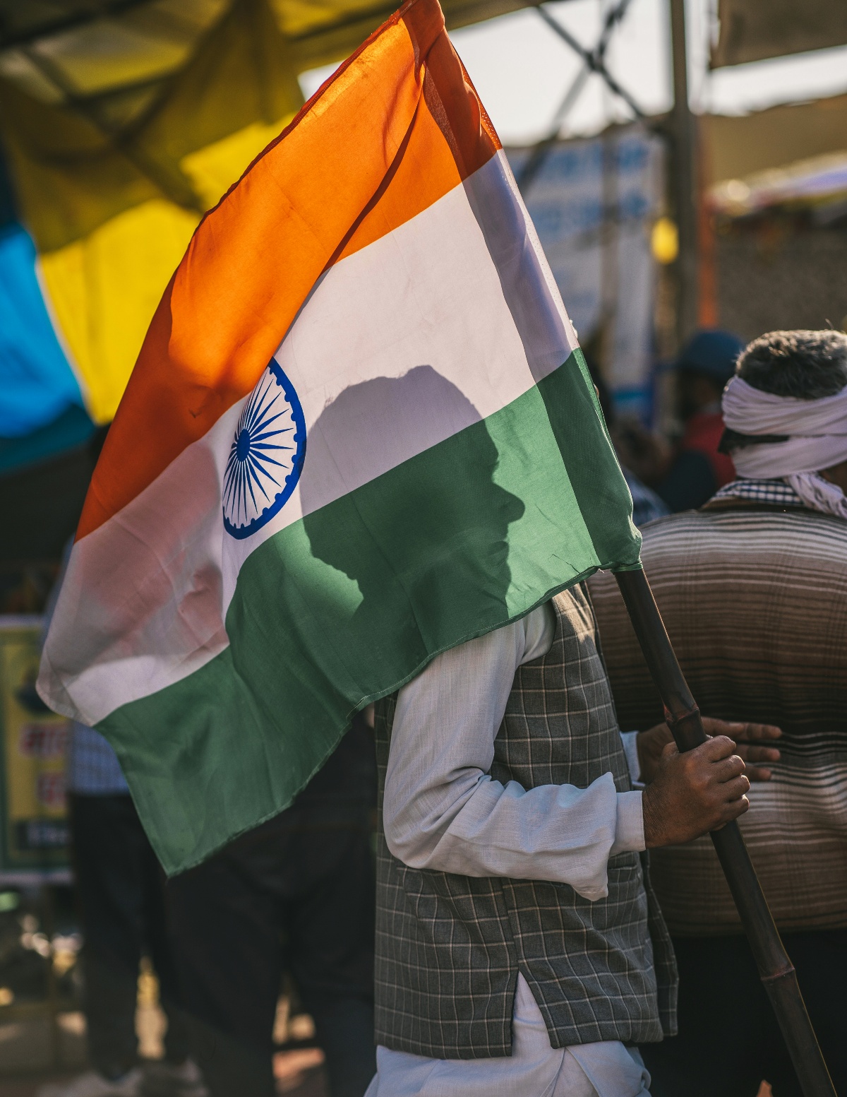 A man in a checked vest carries an Indian tricolor flag over his shoulder, his silhouette visible through the translucent saffron, white, and green fabric with its blue Ashoka Chakra. Behind him, a crowd gathers under a yellow canopy in warm, late-day light.
