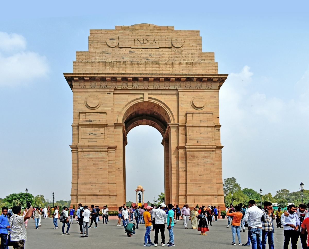 India Gate, a triumphal sandstone arch inscribed with the word "INDIA" and rows of commemorative text, rises against a partly cloudy sky. Crowds of visitors gather and walk across the surrounding plaza, with a small domed canopy and trees visible beneath the arch.