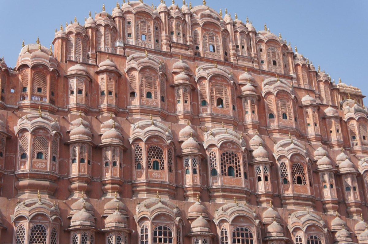 The Hawa Mahal's pink sandstone facade rises in tiered rows of small balconies, each topped with a domed canopy and adorned with intricate latticed windows. The honeycomb-like structure features hundreds of these small openings arranged across multiple stories against a clear blue sky.