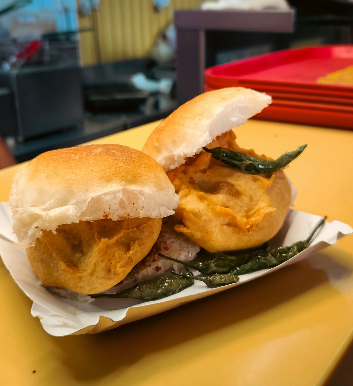 Two vada pav sandwiches, fried potato fritters tucked into soft bread rolls, sit on a paper tray garnished with fried green chilies. The dish rests on a yellow table with a blurred kitchen setup and stacked red trays visible in the background.