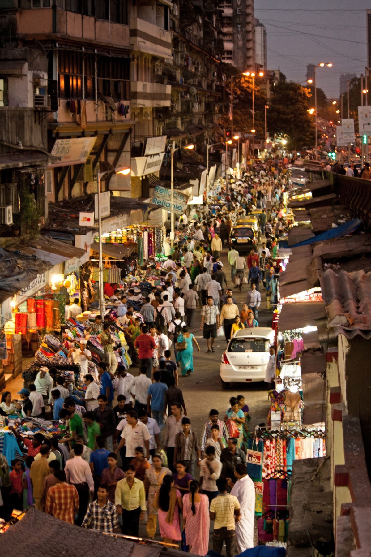 An elevated view shows a crowded Mumbai street market at dusk, with hundreds of pedestrians moving through a narrow road lined with clothing stalls and shops on both sides. Cars navigate slowly through the dense foot traffic as streetlights and shop signs begin to illuminate the scene.