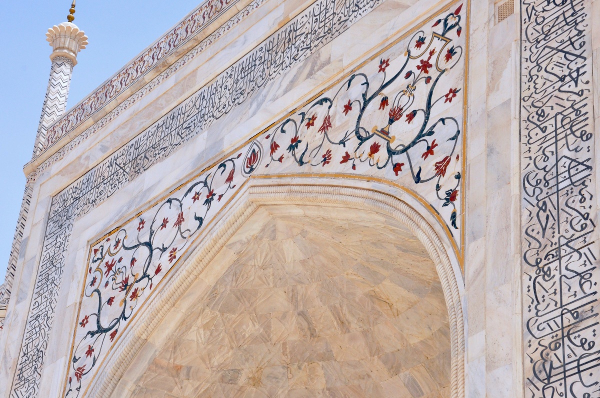 A close-up view of the Taj Mahal's marble archway shows intricate floral inlay work in red and dark green alongside bands of black calligraphic script bordering the arch. A slender minaret with chevron patterning is visible against a clear blue sky in the upper left.