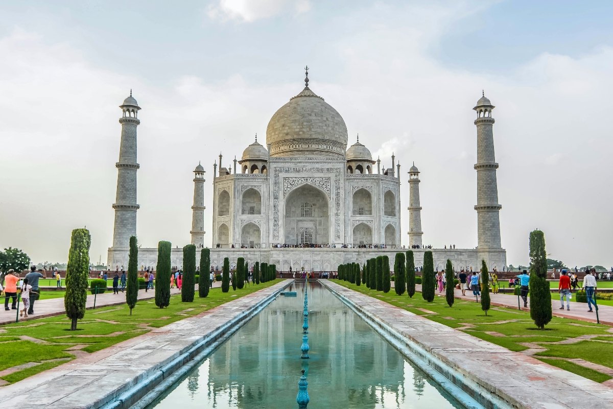 The Taj Mahal's white marble mausoleum, topped by a central dome and flanked by four minarets, is reflected in a long rectangular pool lined with cypress trees and manicured lawns. Visitors walk along the surrounding pathways and gardens beneath an overcast sky.