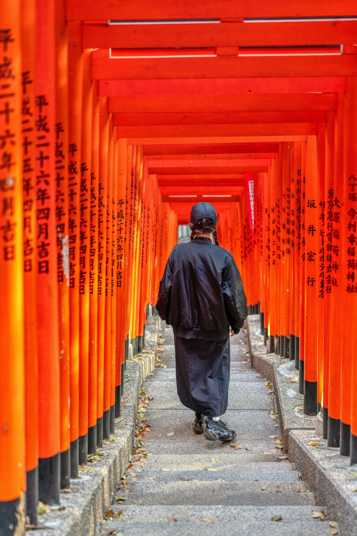 A person dressed in black walks away from the camera up a stone staircase through a tunnel of densely packed vermilion torii gates, each inscribed with black Japanese characters. Scattered autumn leaves dot the steps as the gates recede into the distance, creating a strong sense of depth and repetition.
