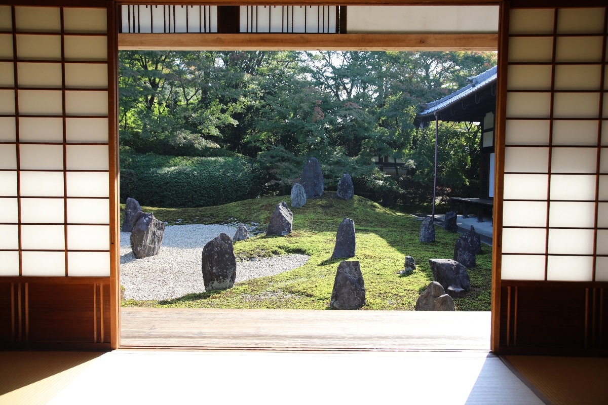 A karesansui (dry landscape) garden is viewed through the open sliding shoji screens of a tatami room, with upright dark stones of varying heights arranged across a ground cover of raked gravel and moss. Dense green trees and trimmed shrubs fill the background, and the tiled eave of an adjacent building is visible at the upper right.