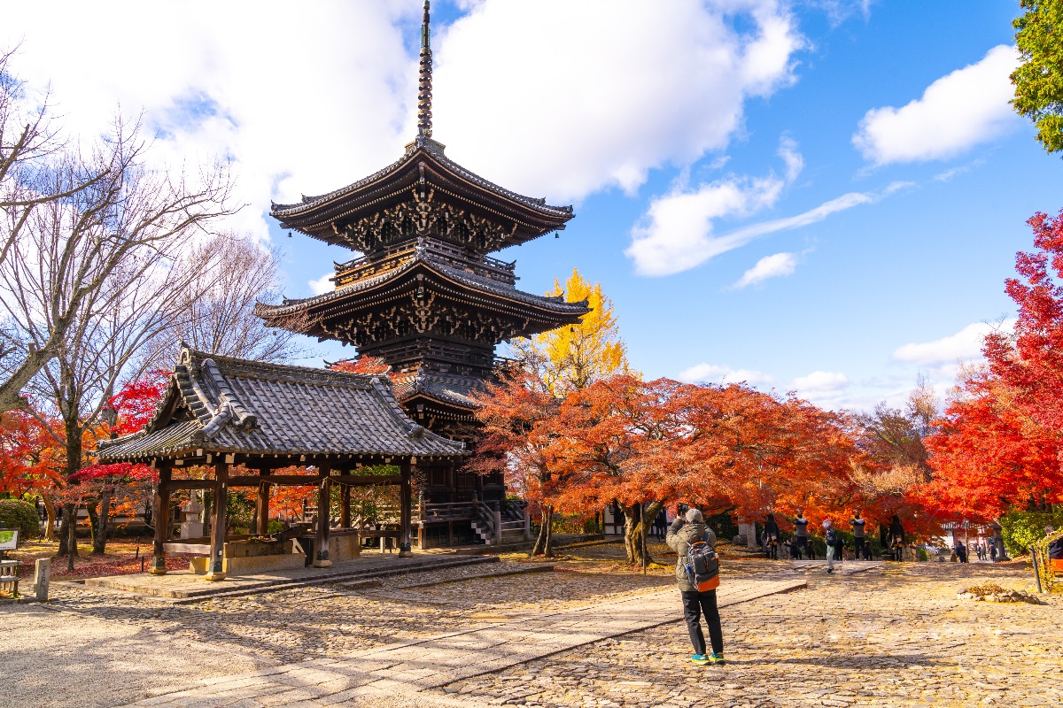 A dark wooden three-story pagoda rises above a covered bell pavilion in a temple courtyard, surrounded by maple trees in peak autumn color ranging from orange and red to a single bright yellow ginkgo. Several visitors photograph the scene from a stone-paved path under a partly cloudy blue sky.