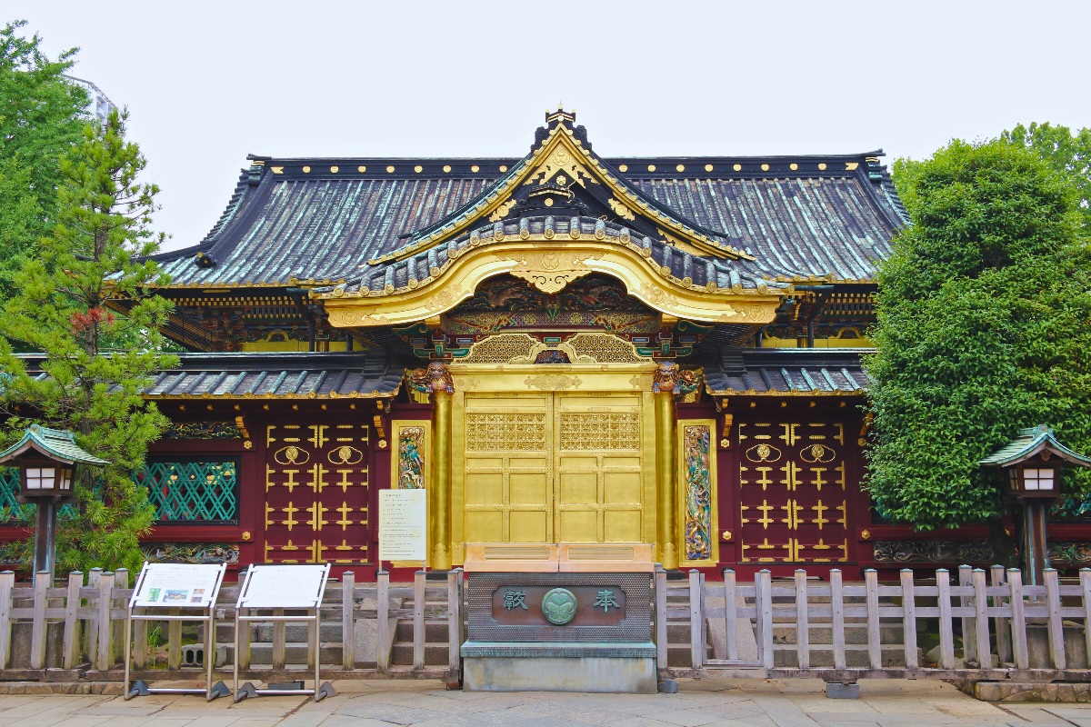 The ornate facade of a Japanese shrine building features a large gilded central gate with intricate carved relief work, set against deep red lacquered walls decorated with gold medallions and geometric patterns beneath a sweeping black tiled roof. Stone lanterns and a low wooden fence flank the entrance, with green trees framing either side of the structure under an overcast sky.