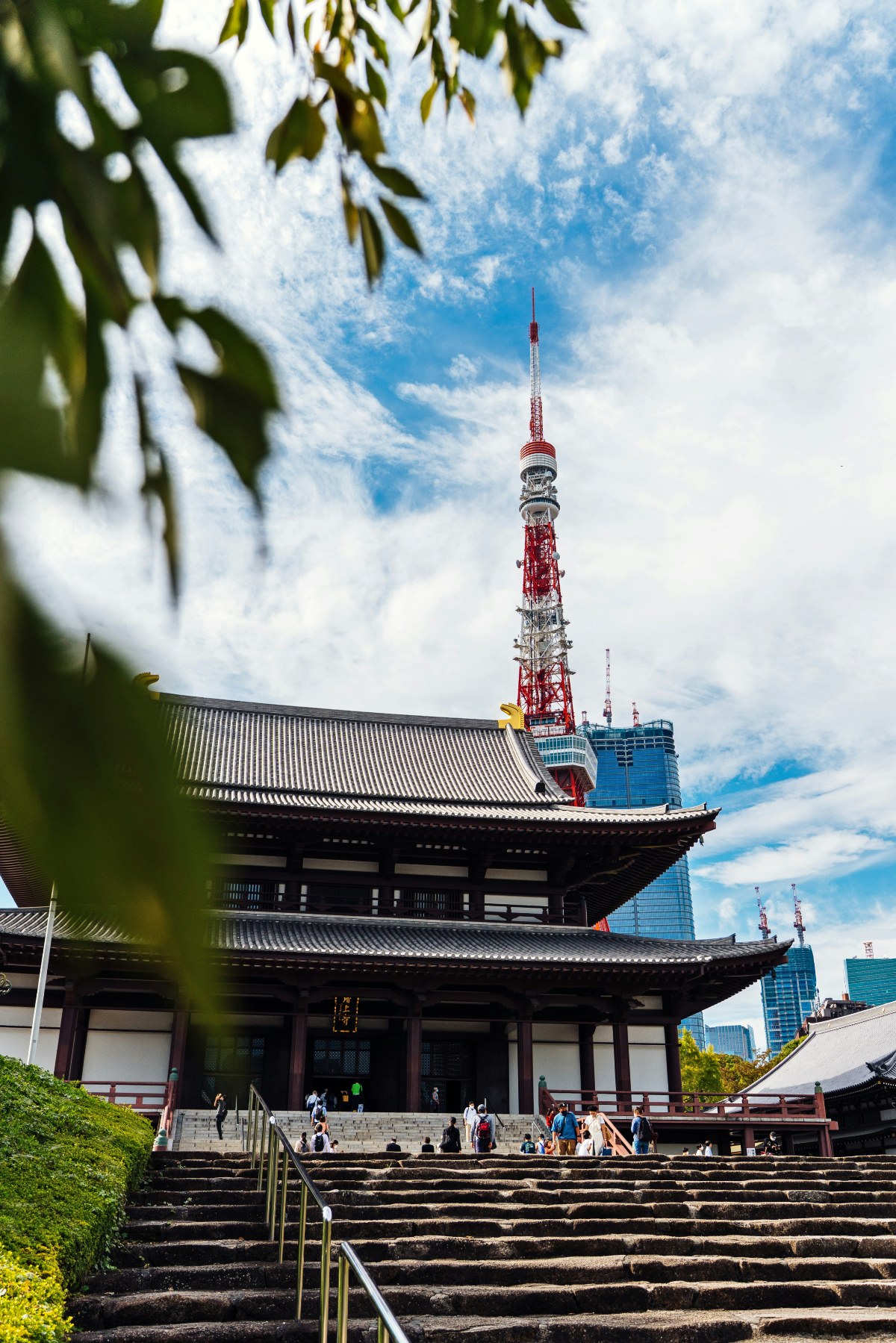 The main hall of Zojo-ji Temple, a traditional Japanese Buddhist structure with sweeping tiled roofs and dark wooden columns, is viewed from the base of a wide stone staircase where visitors gather. Tokyo Tower's red-and-white lattice frame rises directly behind the temple against a partly cloudy sky, with modern glass skyscrapers visible to the right.