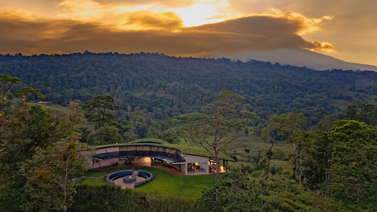 Aerial view of a curved, low-profile lodge building with a living green roof set in a clearing surrounded by dense forest, with a circular fire pit terrace in front of it. A large volcano partially obscured by clouds rises in the background under an orange and gray sunset sky.