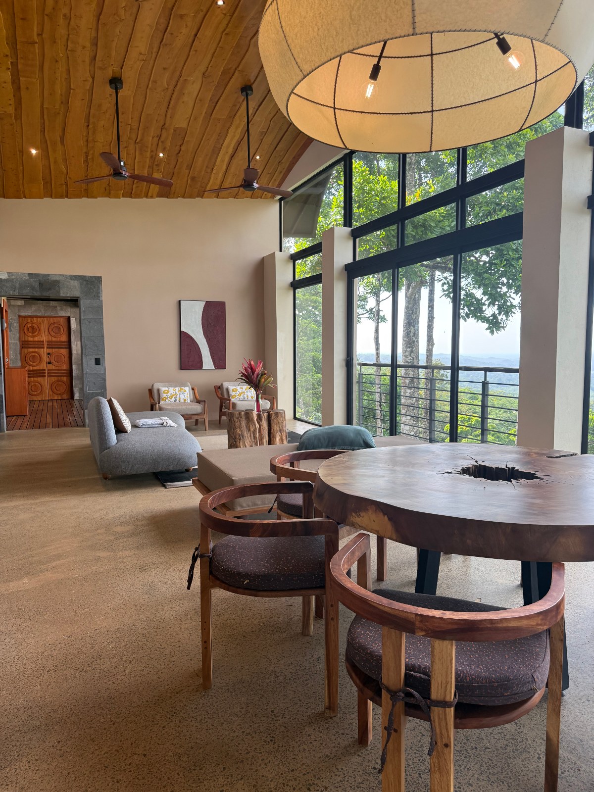 A vaulted wood-ceilinged living and dining area features a round live-edge wood table with wooden chairs in the foreground, gray sofas, and tree-stump side tables, with a large woven pendant lamp hanging overhead. Floor-to-ceiling windows on the right reveal a forested hillside view, while wooden double doors and stone trim are visible at the far end of the room.