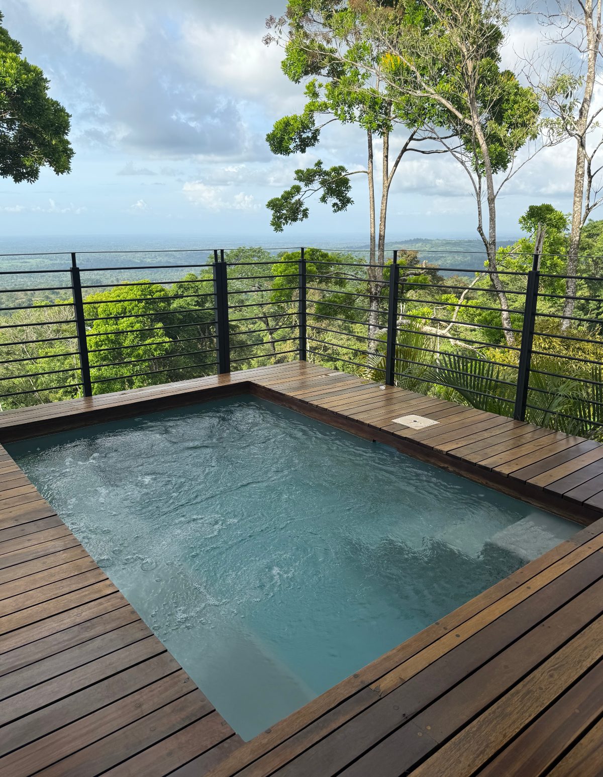 A small rectangular plunge pool with rippling water sits on a wooden deck enclosed by a black metal railing, perched at the edge of a forested hillside. Trees frame the view beyond the railing, with a hazy expanse of lowland forest visible under a partly cloudy sky.