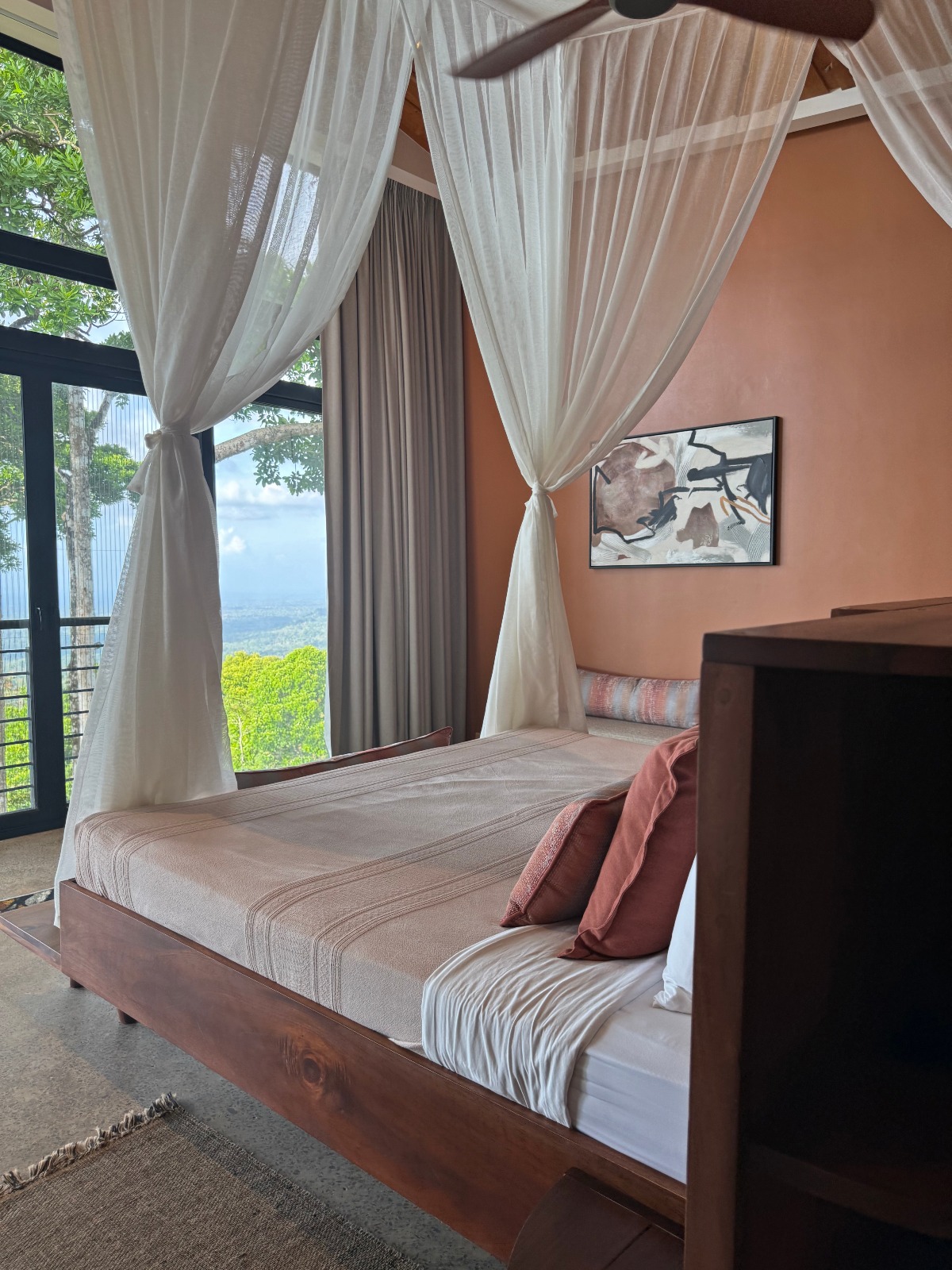 A bedroom with a wooden four-poster bed draped in sheer white curtains tied back at the corners, set against a terracotta-colored wall with an abstract framed artwork. Floor-to-ceiling windows to the left look out over a forested valley below.