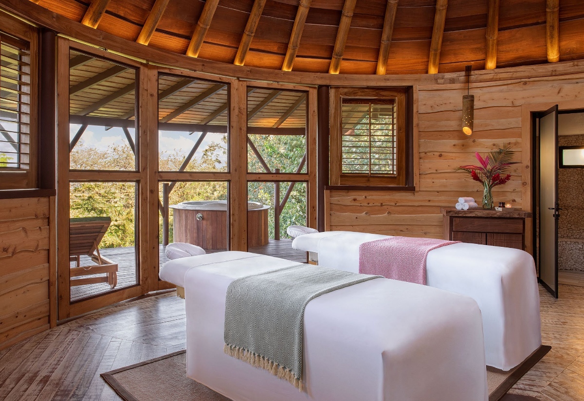 A round, thatched-roof spa pavilion with two side-by-side massage tables draped in white linens and colored throw blankets, surrounded by wood-paneled walls and large windows. A wooden soaking tub and lounge chair sit on an outdoor deck visible through the windows, with forested hills in the background.