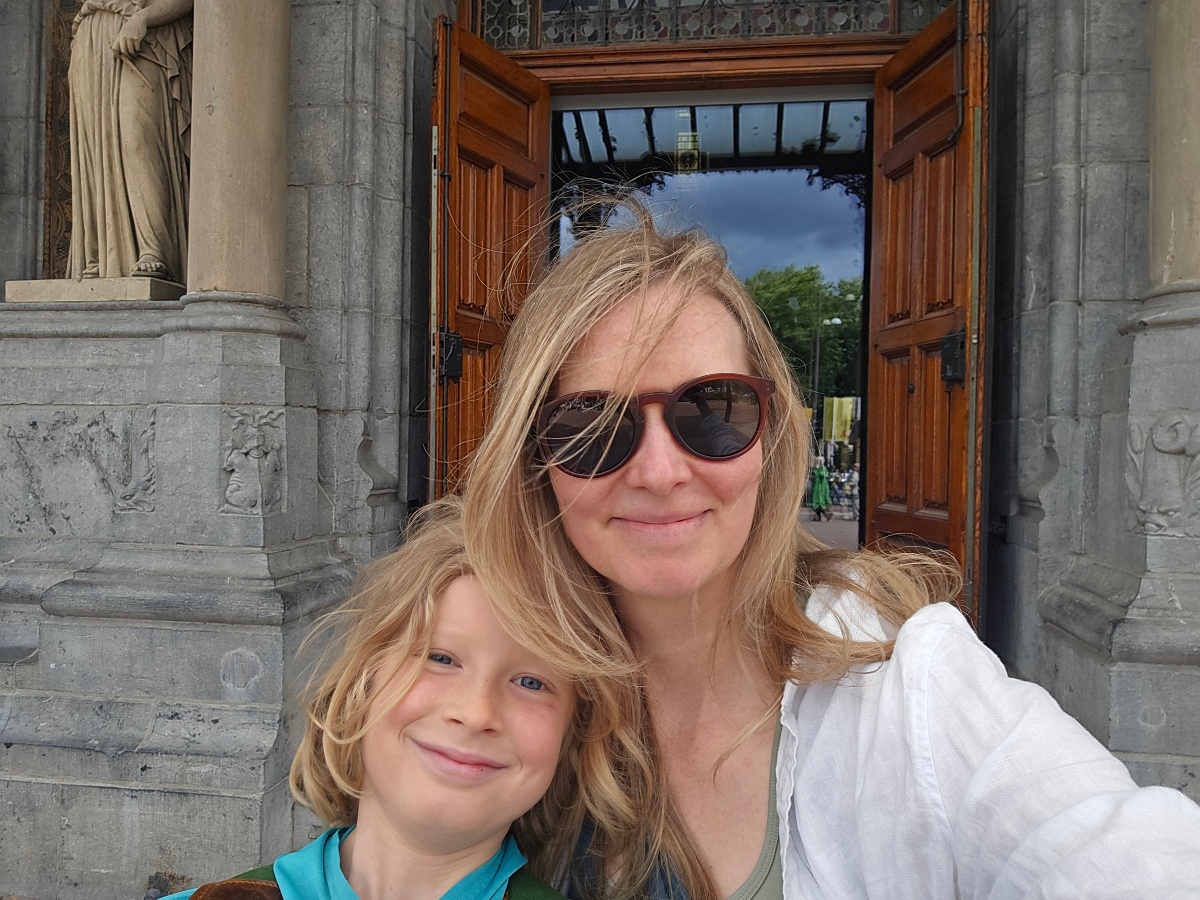 A woman in sunglasses and a white blouse takes a close-up selfie with a smiling blond child in a teal shirt leaning against her shoulder. They stand in the arched stone entryway of an ornate historic building, with tall wooden doors open behind them revealing a tree-lined street beyond.