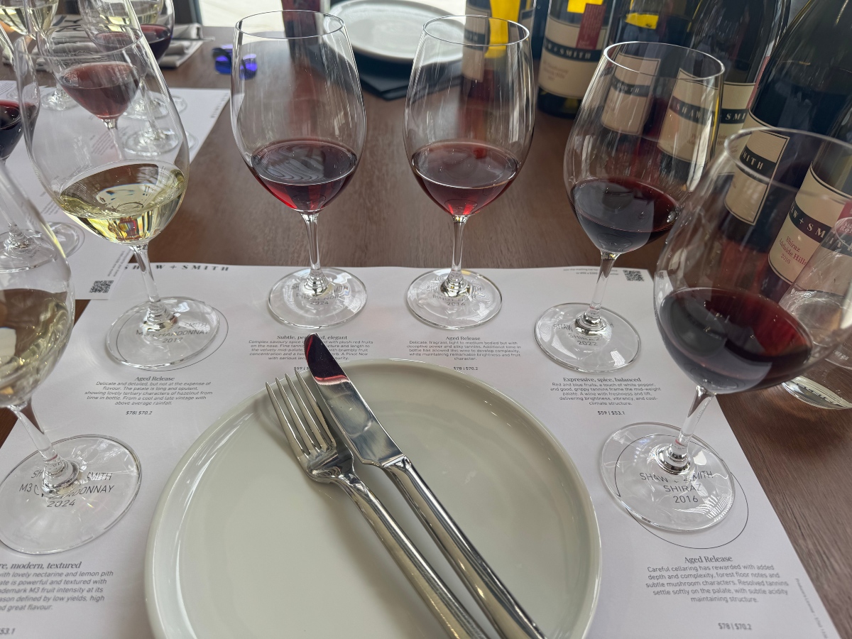 A tasting flight of white and red wines is arranged on a printed placemat at Shaw + Smith winery, with each glass positioned over its label and tasting notes. A white plate with a fork and knife sits in the foreground, and wine bottles line the back of the wooden table.