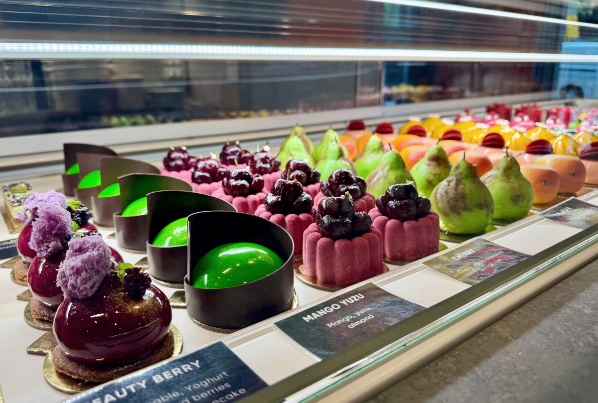 Rows of sculptural French pastries fill a glass display case, including glossy burgundy domes, bright green mango yuzu desserts wrapped in chocolate, and cakes shaped like realistic pears and cherries. Printed labels identify flavors such as Beauty Berry, Mango Yuzu, and Cherry.