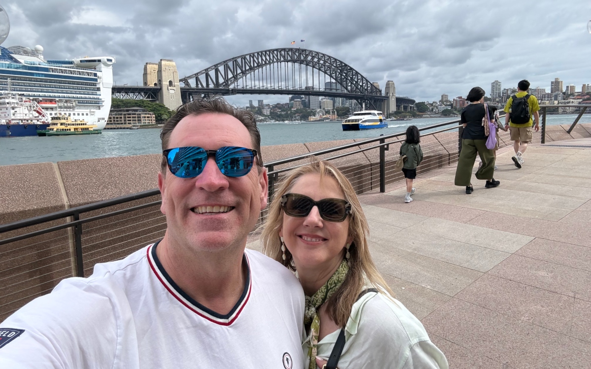 A smiling couple in sunglasses takes a selfie along a waterfront walkway with the Sydney Harbour Bridge spanning the background. A cruise ship, ferries, and pedestrians are visible along the harbor under a cloudy sky.