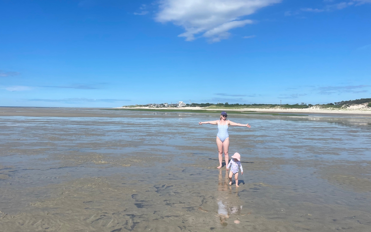 A woman in a light blue swimsuit stands with arms outstretched on wide tidal flats at low tide, with a toddler in a pink sun hat walking ahead of her through shallow water. Sand dunes, beach grass, and distant houses line the shoreline beneath a bright blue sky.