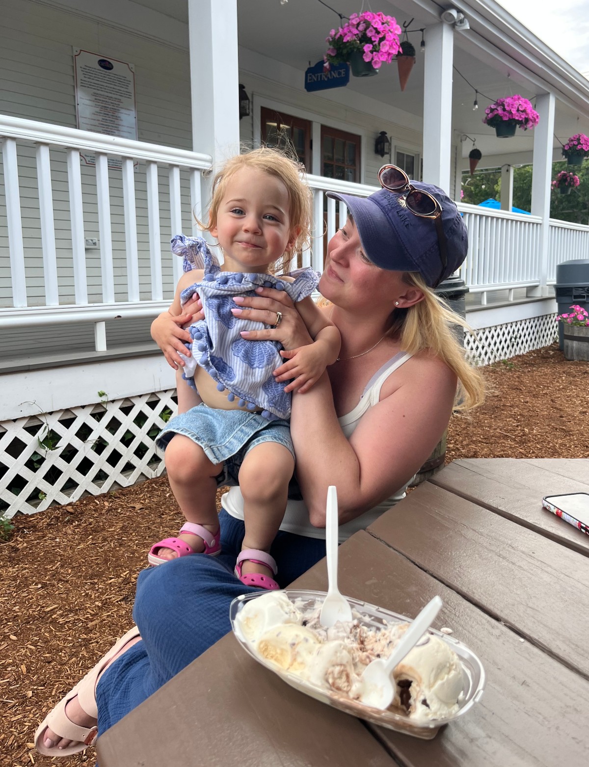 A woman in a blue baseball cap holds a smiling toddler in a blue-and-white outfit on her lap at a picnic table, with a shared dish of ice cream and plastic spoons in the foreground. Behind them is a white ice cream shop porch with railings, hanging baskets of pink flowers, and an entrance sign.