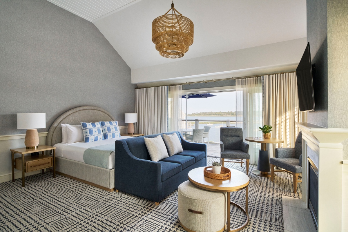 A coastal-style hotel room with a king bed, navy blue sofa, gray armchairs, and a woven rattan pendant light under a vaulted ceiling. Sliding glass doors open to a private balcony with umbrella-shaded seating overlooking the water.