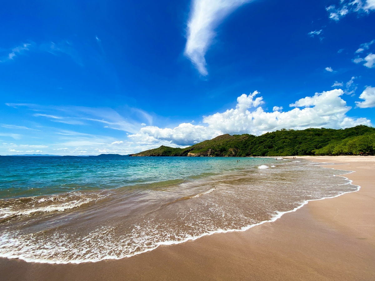 A sandy beach curves along the right side of the frame, where shallow turquoise water rolls ashore in gentle, foamy waves under a bright blue sky scattered with white clouds. In the background, a green, tree-covered headland juts into the sea, and a few people can be seen wading in the distant shallows.