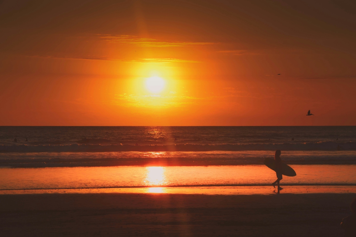 The sun hangs low above the ocean horizon, casting a vivid orange glow across the sky and a bright reflection on the wet sand below. A silhouetted person carrying a surfboard walks along the shoreline at right, while a bird flies overhead and a few small figures dot the waves in the distance.