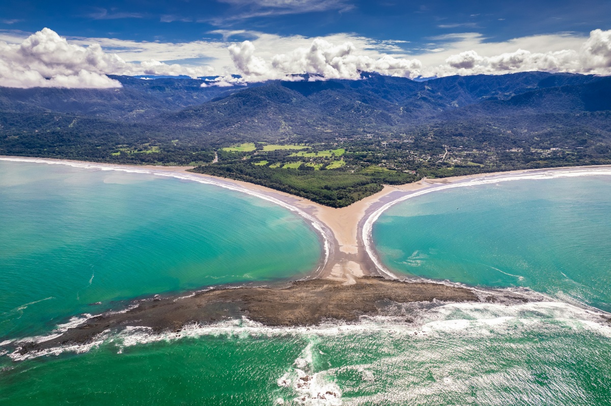 An aerial view shows a narrow sandbar extending from a forested coastline into turquoise water, where it meets a wide, dark rocky outcrop, together forming a shape resembling a whale's tail. Beyond the shore, green fields and scattered buildings give way to forested mountains topped with white clouds under a blue sky.