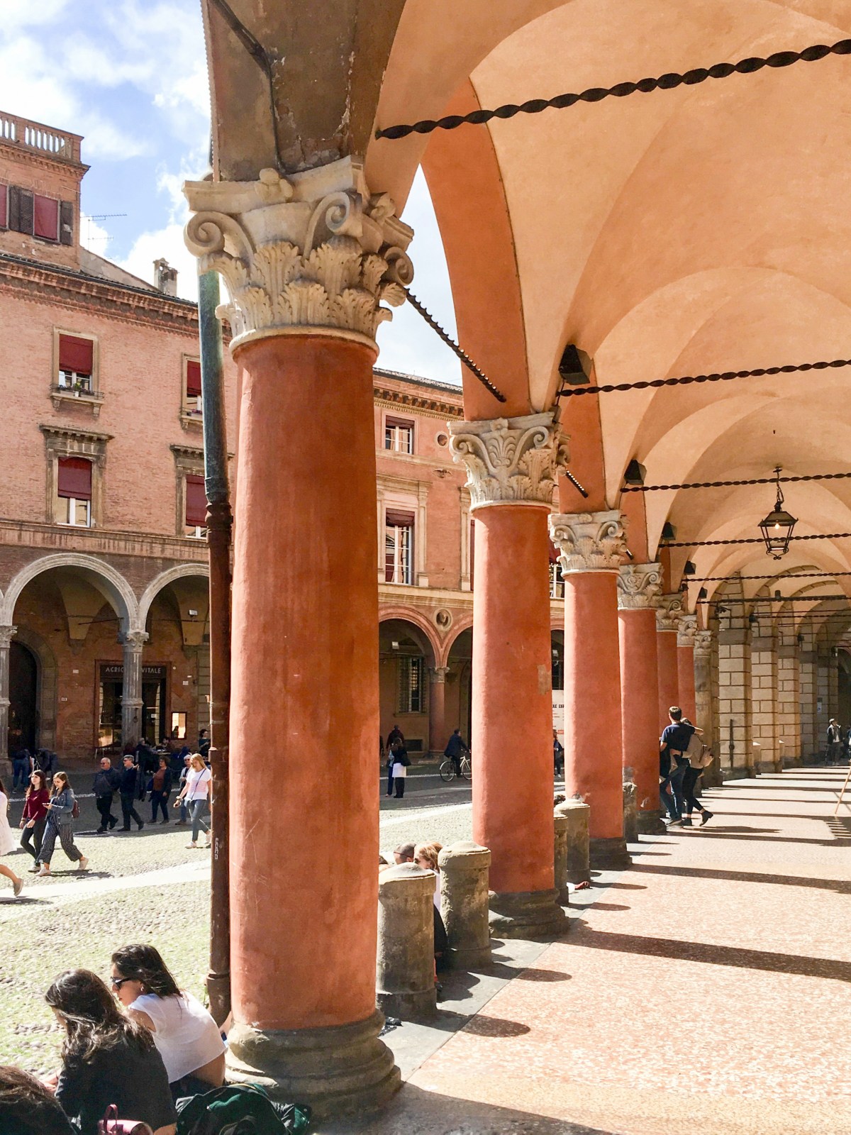 A row of terracotta-colored columns with carved Corinthian capitals supports the vaulted arches of a portico in Bologna, with a lantern hanging from the ceiling. Pedestrians walk along the sunlit cobblestone square beyond, where more arcaded brick buildings with red-shuttered windows line the piazza.
