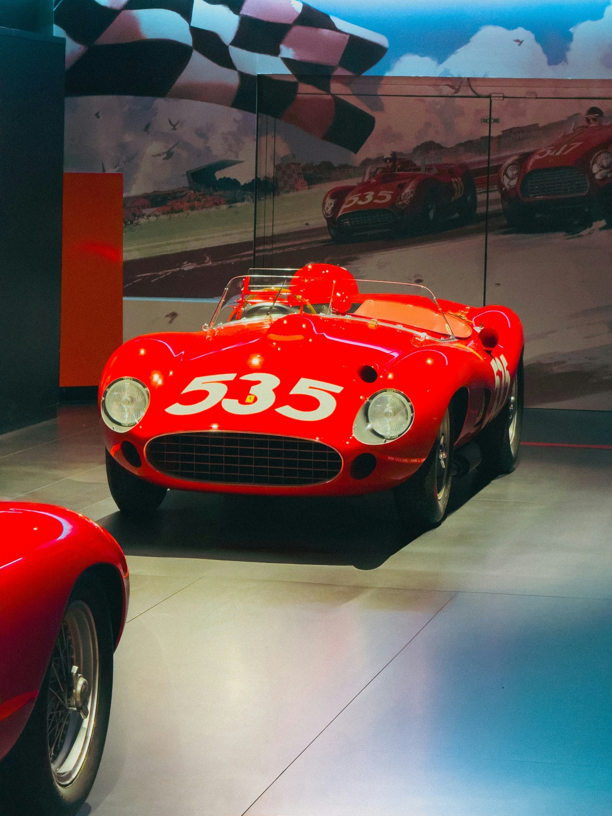 A vintage red Ferrari race car with the number 535 painted in white on its hood is displayed in a museum gallery, its open cockpit fitted with a low windscreen. Behind it, a large mural depicts the same car racing under a checkered flag, and the nose of a second red Ferrari appears in the foreground.
