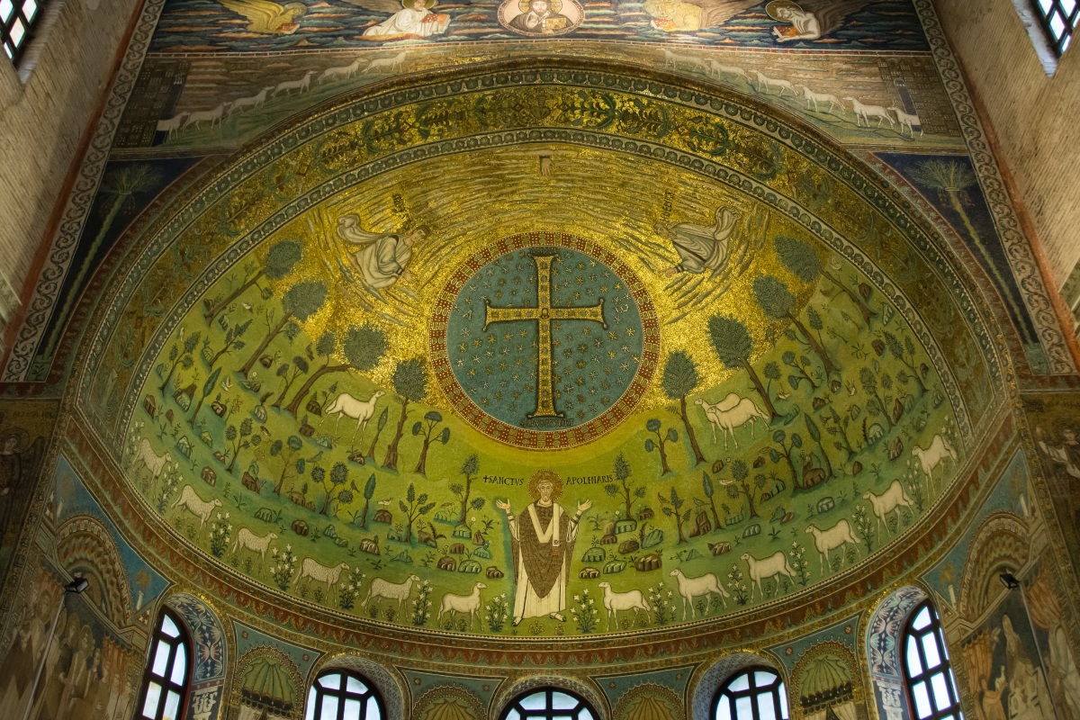 The apse mosaic of the Basilica di Sant'Apollinare in Classe depicts a jeweled gold cross inside a star-filled blue medallion, set against a green landscape of trees, rocks, and white sheep. Below the cross, Saint Apollinaris stands with arms raised in prayer, flanked by lambs, while figures and sheep line the golden band above the arch.