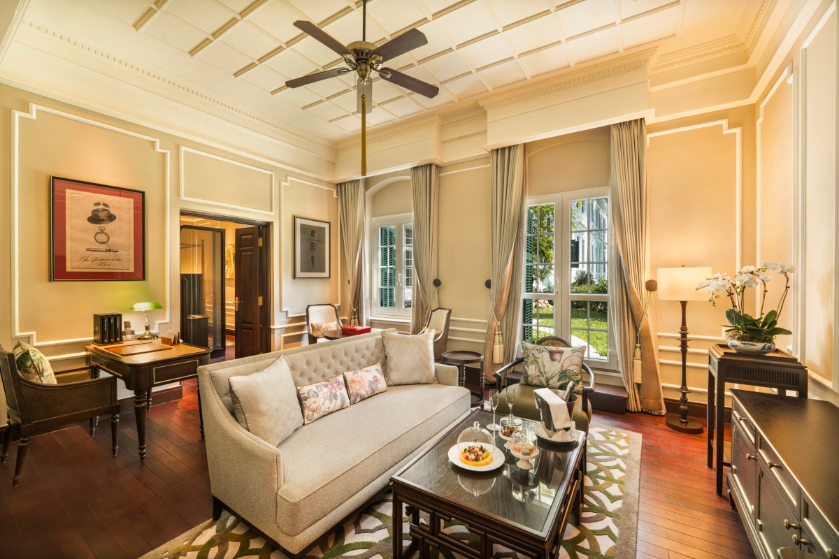The Somerset Maugham Suite at the Sofitel Legend Metropole Hanoi features cream paneled walls, a coffered ceiling with a fan, and tall shuttered windows draped in silk curtains. A tufted beige sofa faces a dark wood coffee table set with refreshments, with a writing desk and orchid arrangement nearby.