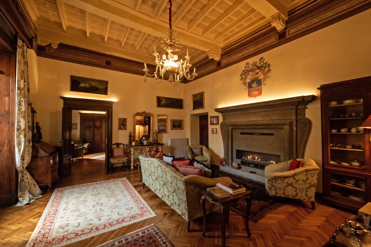 A sitting room at Palazzo Misciattelli features a coffered ceiling, crystal chandelier, and a large stone fireplace topped with a painted coat of arms. Damask upholstered sofas and armchairs surround the lit fireplace, with antique furniture, framed paintings, and Oriental rugs on the herringbone wood floor.