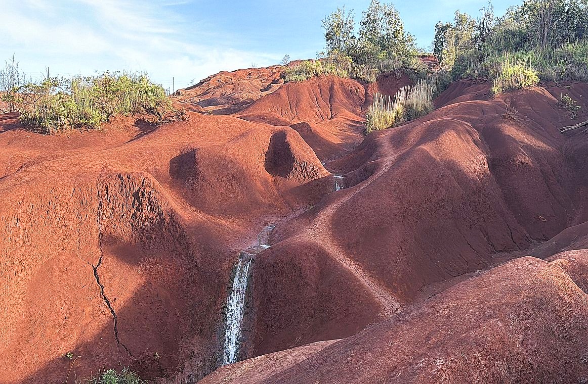 A narrow waterfall streams down a channel carved into steep red dirt slopes at Red Dirt Falls in Waimea Canyon State Park on Kauai. Green shrubs and grasses grow along the top edges of the eroded rust-colored ridges, with a pale blue sky and thin clouds above.