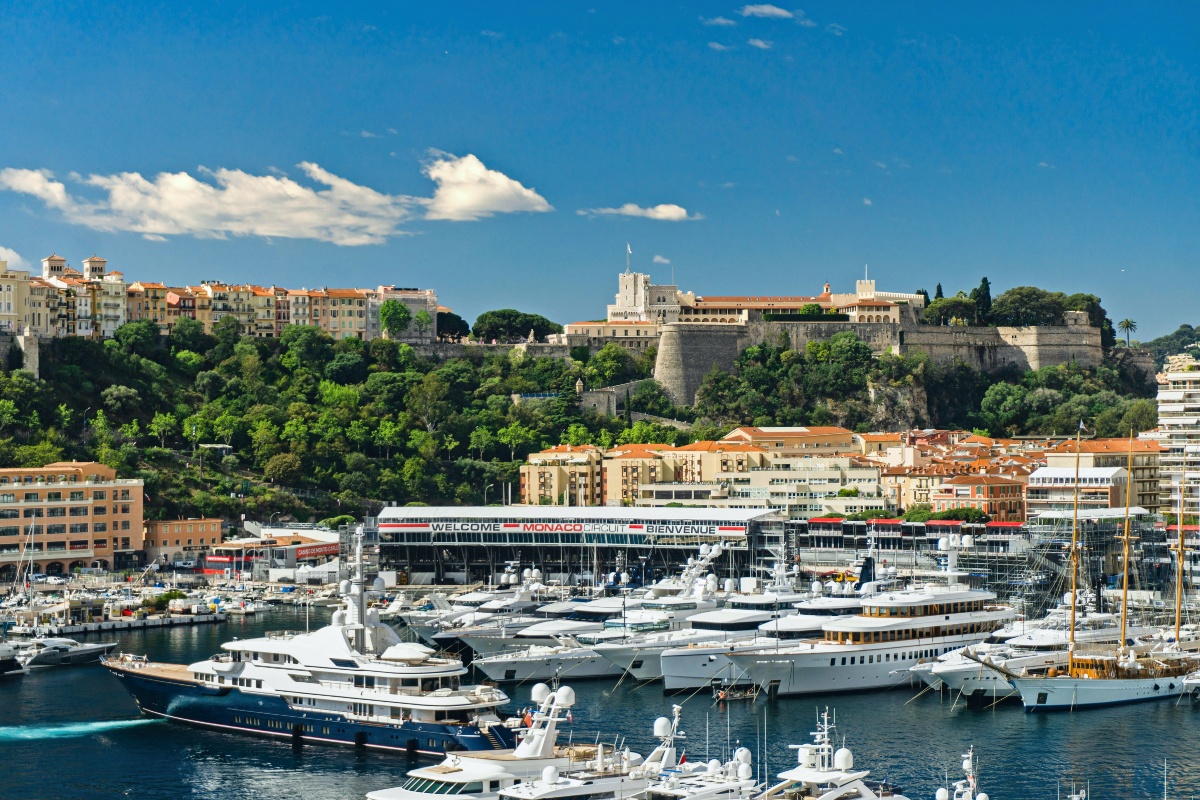 Rows of large white yachts fill Monaco's Port Hercule, with a grandstand structure along the waterfront marked with signage for the Monaco circuit. Above the harbor, pastel buildings with terracotta roofs and a fortified palace complex sit atop a green, wooded rock outcrop under a blue sky with scattered clouds.