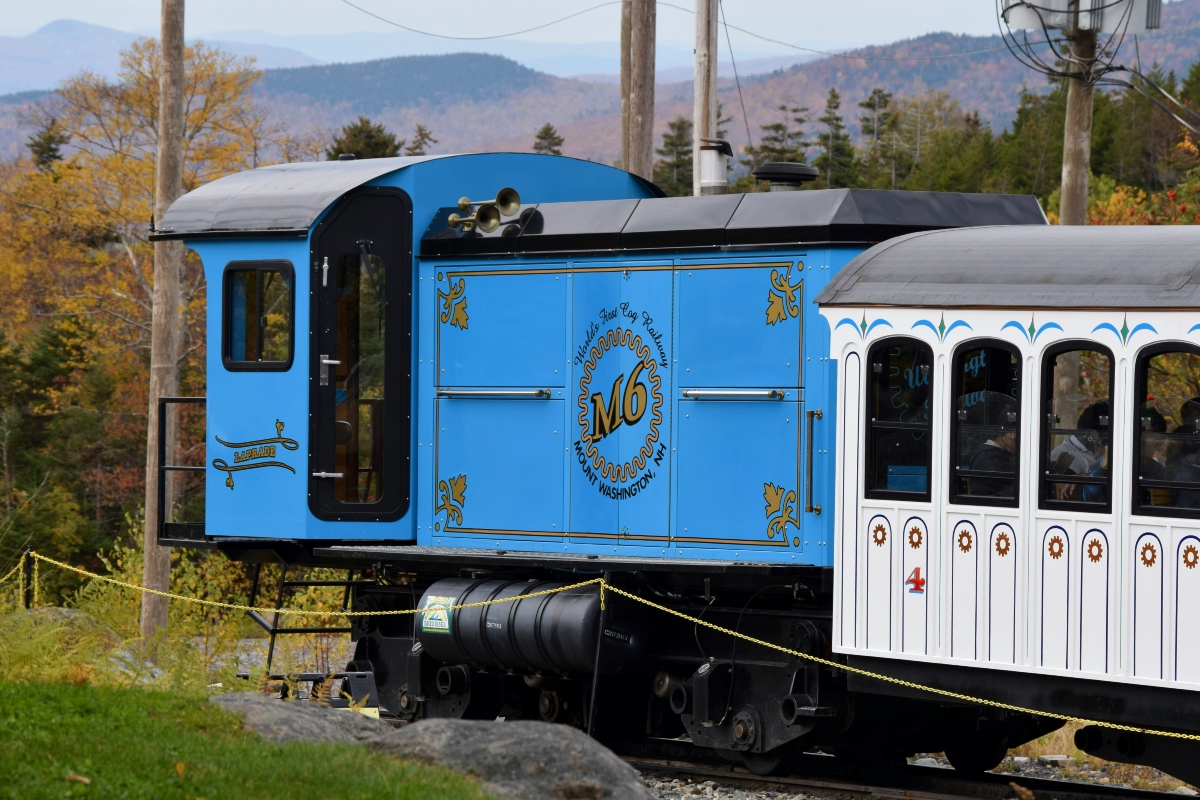 A bright blue Mount Washington Cog Railway locomotive marked "M6" with gold scrollwork sits on the track, coupled to a white passenger car with arched windows and seated riders. Fall foliage in yellow and orange fills the trees behind it, with layered mountain ridges visible in the distance.