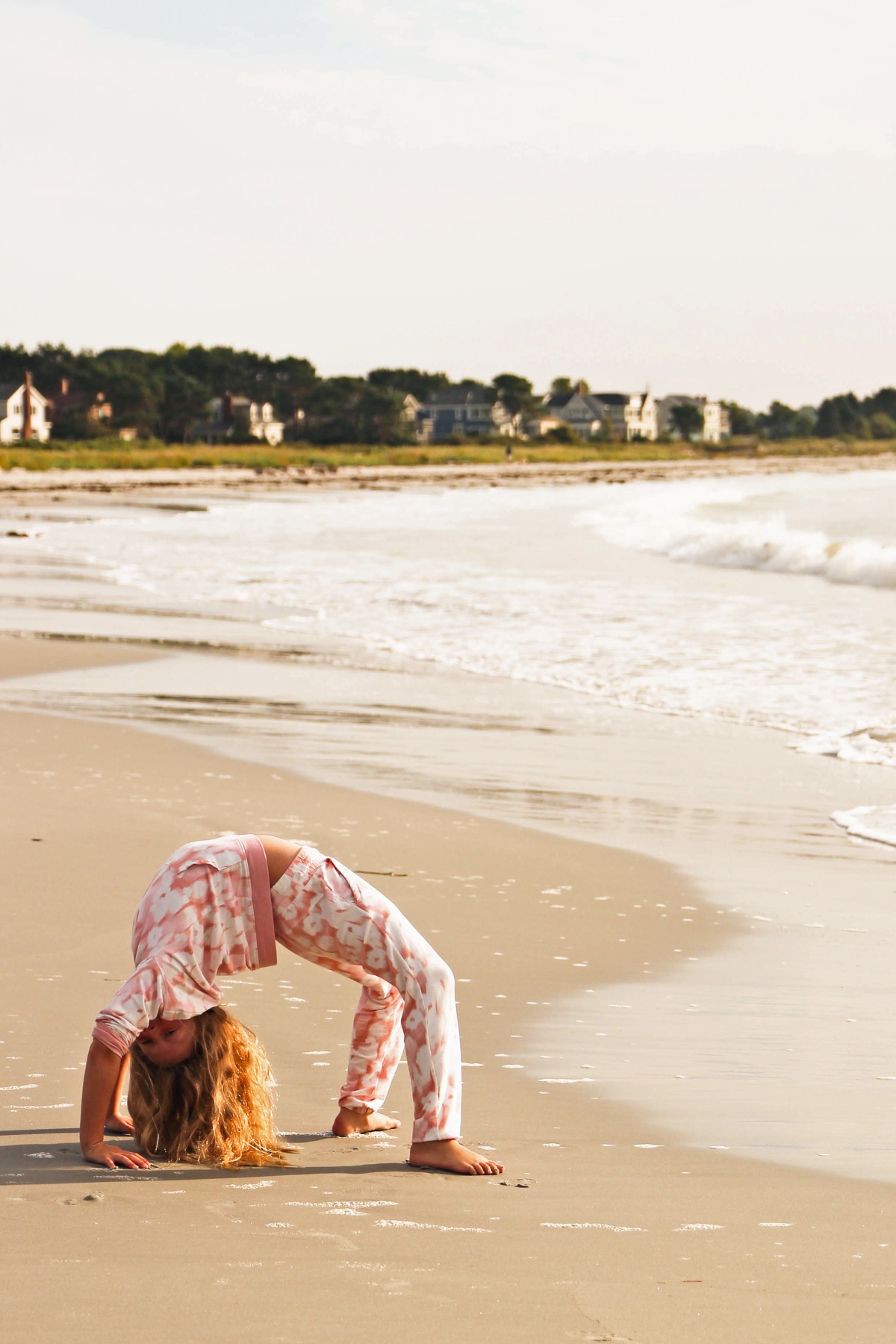 A young girl in a pink and white tie-dye outfit does a backbend barefoot on wet sand near the water's edge. Behind her, small waves break along a wide, empty beach, with shingled houses and a tree line visible along the shore under a hazy sky.