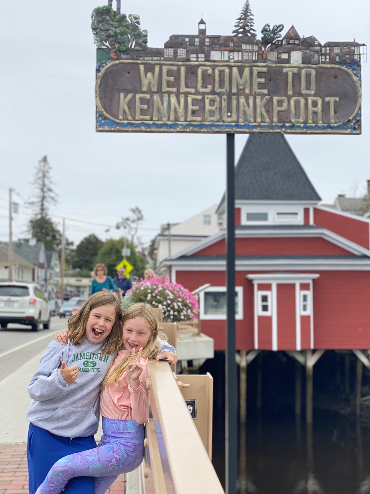 Two young girls pose together at a bridge railing beneath a decorative metal "Welcome to Kennebunkport" sign, one giving a thumbs-up and the other a peace sign. Behind them, a red shingled building on pilings stands over the water, with a street, parked cars, and flower boxes visible along the left side under an overcast sky.