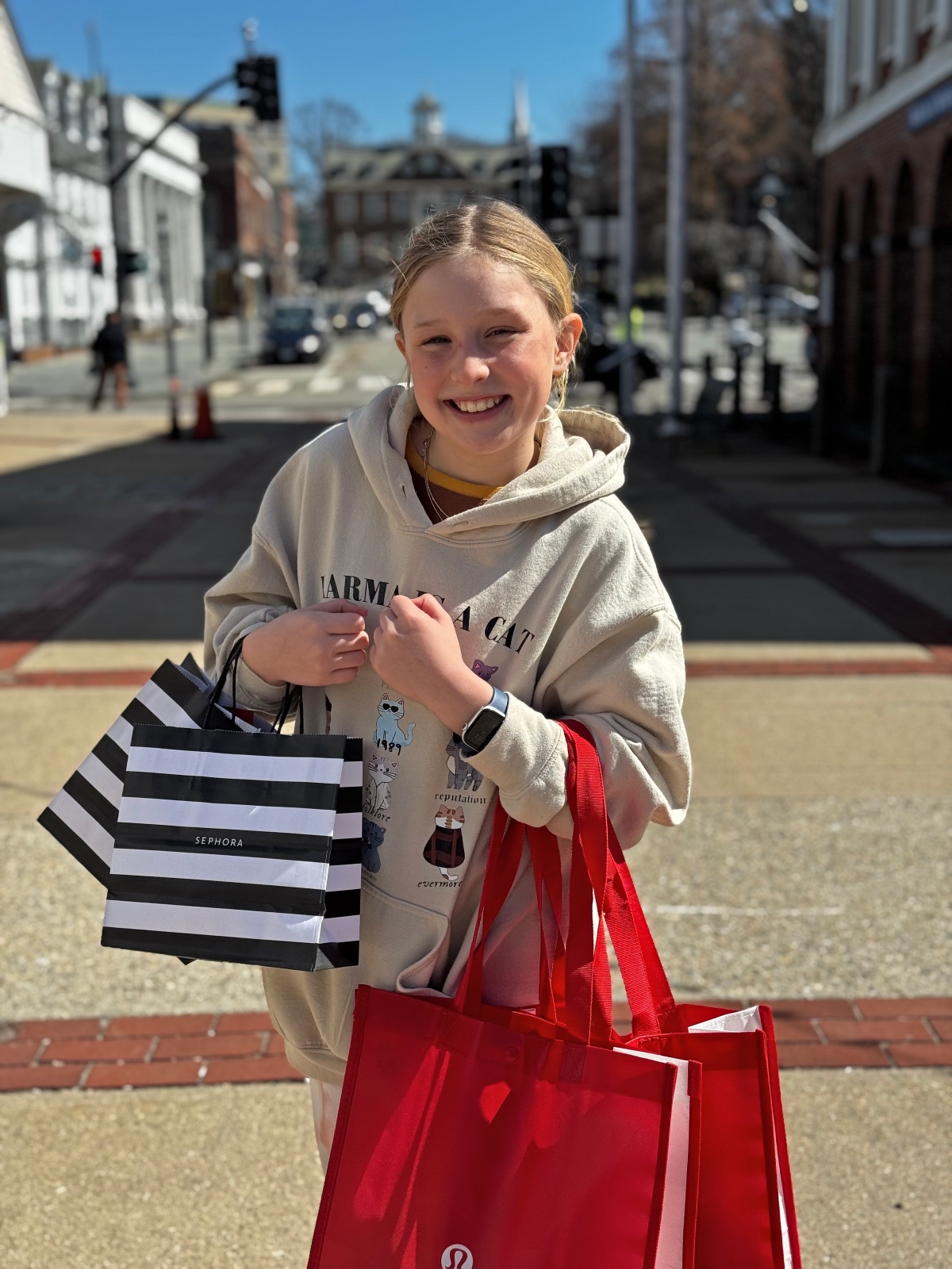 A smiling girl in a cream graphic hoodie stands on a sidewalk holding several shopping bags, including a black-and-white striped bag and a red reusable tote. Behind her, a sunlit street corner with brick and clapboard buildings, traffic lights, and a crosswalk stretches into the background under a clear blue sky.