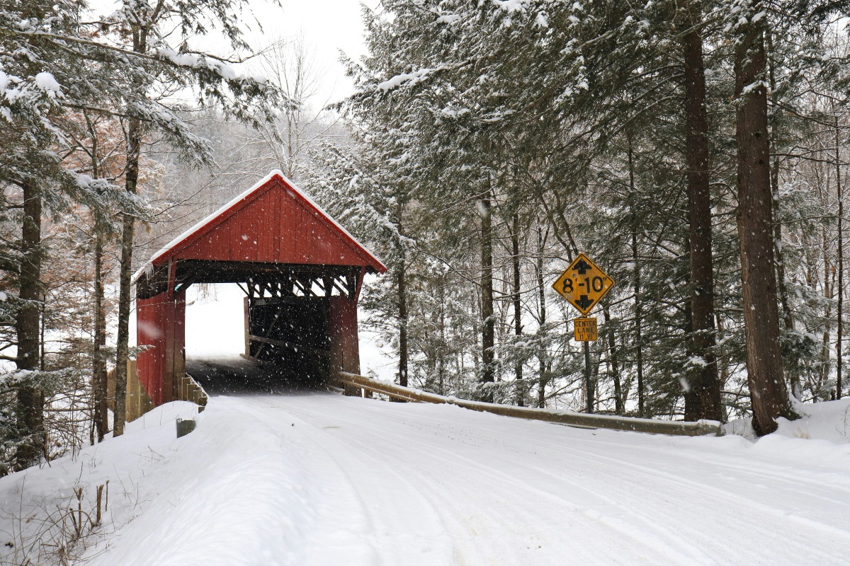A red covered bridge with a single lane sits at the end of a snow-covered road, framed by evergreens and bare trees heavy with fresh snow. A yellow clearance sign reading 8'-10" stands at the roadside on the right as snow falls.