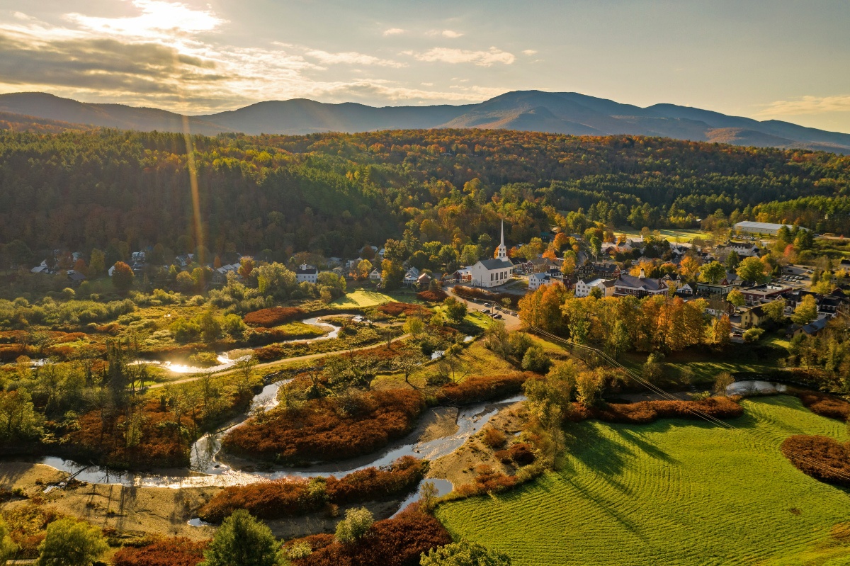 An aerial view of a small New England village centered on a white church with a tall steeple, surrounded by clustered buildings and trees in fall color. A winding stream cuts through open meadows in the foreground, with forested hills and a mountain ridge rising behind the town under a hazy sunlit sky.